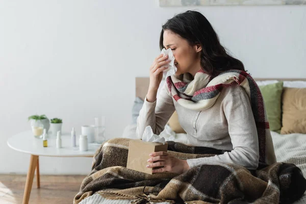 暖かいスカーフの病気の女性が毛布の下に座って紙ナプキンでくしゃみをする — ストック写真