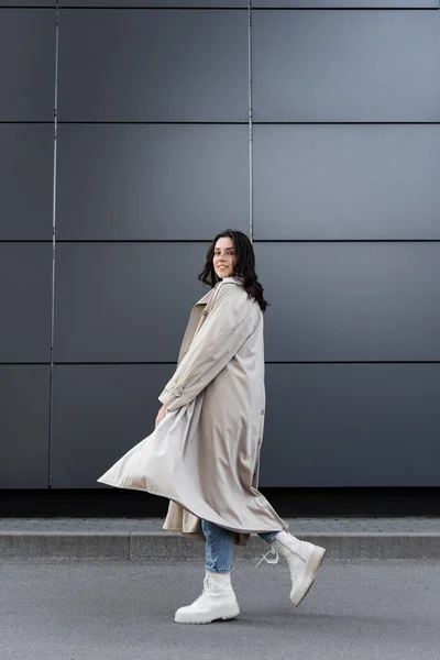 young woman in long raincoat and white leather boots walking along grey wall