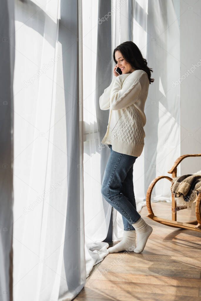 full length view of woman in knitted cardigan and warm socks talking on cellphone near window