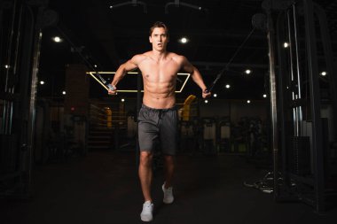 Uzun boylu, şortsuz bir adamın spor salonunda halat çekme egzersiz makinesinde antrenman yaptığını.