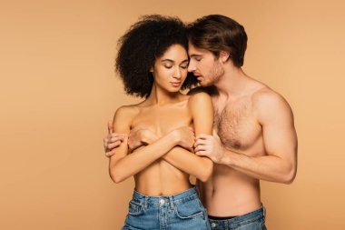 Seksi adam üstsüz İspanyol kadını kucaklıyor. Bej rengi kollarıyla göğsünü kapatıyor.