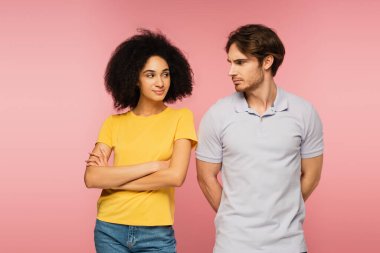positive latin woman and offended man looking at each other isolated on pink clipart