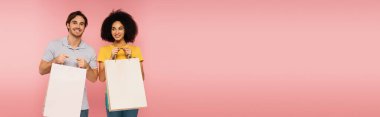 Pembe pankartta izole edilmiş alışveriş torbalarıyla ayakta duran neşeli çoklu etnik çift