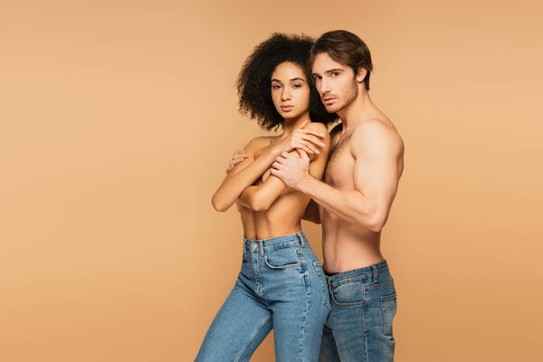 sensual interracial couple in jeans only looking at camera isolated on beige