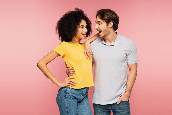 amazed man looking at joyful latin girlfriend standing with hand on hip isolated on pink
