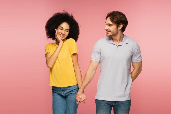 shy hispanic woman holding hands with smiling man hiding something behind back isolated on pink