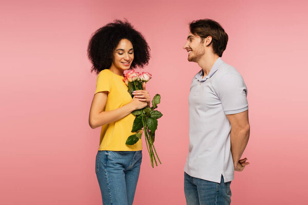 pleased hispanic woman holding bouquet of roses near young boyfriend isolated on pink