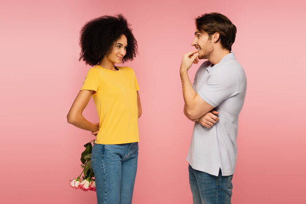 curious man looking at smiling latin girlfriend holding bouquet behind back isolated on pink