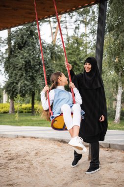 Salıncakta sallanan kızın yanında duran gülümseyen Müslüman anne. 