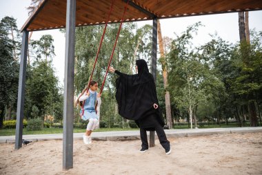 Müslüman anne parkta salıncakta sallanan çocuğun yanında duruyor 