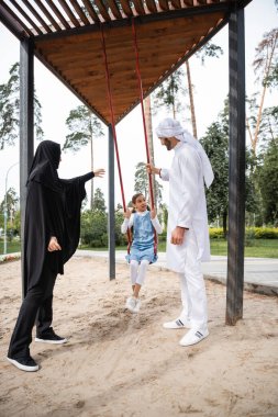 Arabian parents in traditional clothes standing near daughter on swing in park  clipart