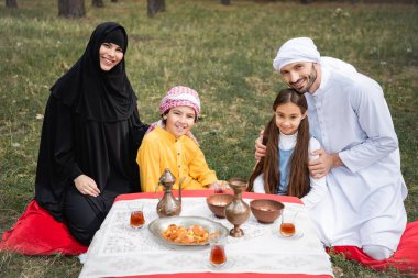 Gülümseyen Arap aileler parkta yemek ve çayın yanında çocuklara sarılıyorlar. 
