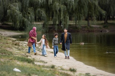 Göl kenarında yürürken el ele tutuşan pozitif Arap ailesi 