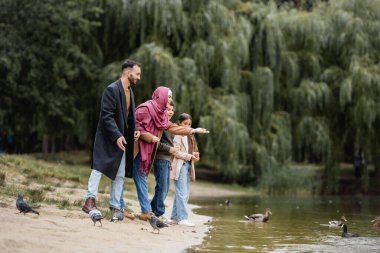 Gülümseyen Müslüman anne göl kıyısındaki kuşlara ve sahildeki çocuklara bakıyor.