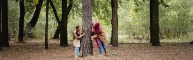 Smiling arabian girl playing with mother near tree in park, banner  clipart