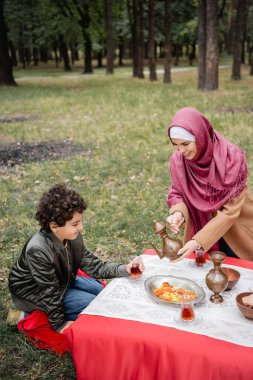 Smiling arabian woman pouring tea near son and dried fruits in park  clipart