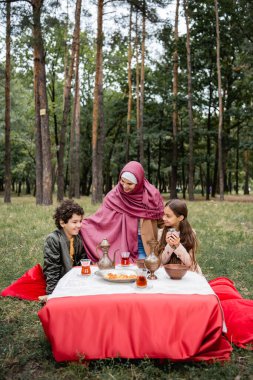 Positive arabian mother hugging kids near tea and dried fruits in park  clipart