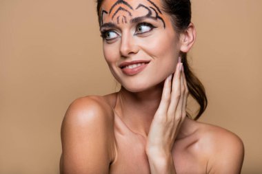 Çıplak omuzlu ve kaplan makyajlı neşeli bir kadın bej rengi bir elbisenin üzerine bakıyor.