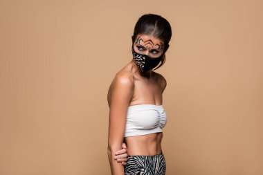 woman with tiger makeup and animal print protective mask looking away isolated on beige clipart