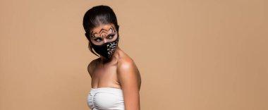 woman with tiger makeup and animal print protective mask looking away isolated on beige, banner clipart