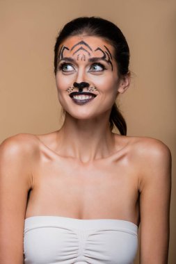 positive woman with bare shoulders and tiger makeup posing isolated on beige clipart