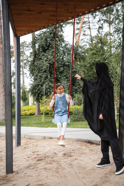 Smiling arabian girl on swing looking at mother in hijab on playground 
