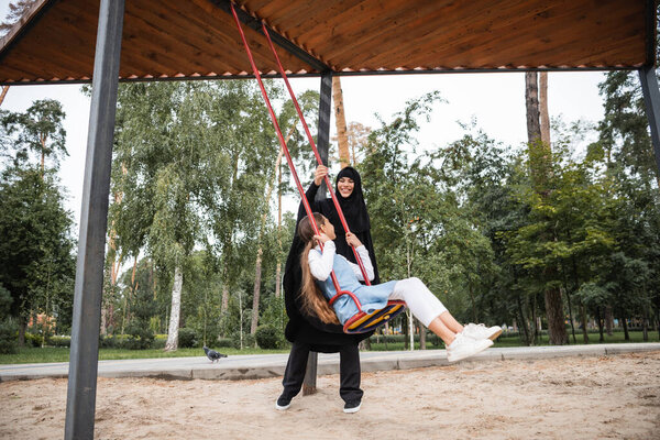 Cheerful muslim mother in hijab standing near daughter on swing in park 