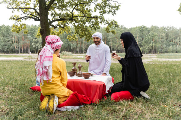 Cheerful arabian family in traditional clothes holding tea in park 