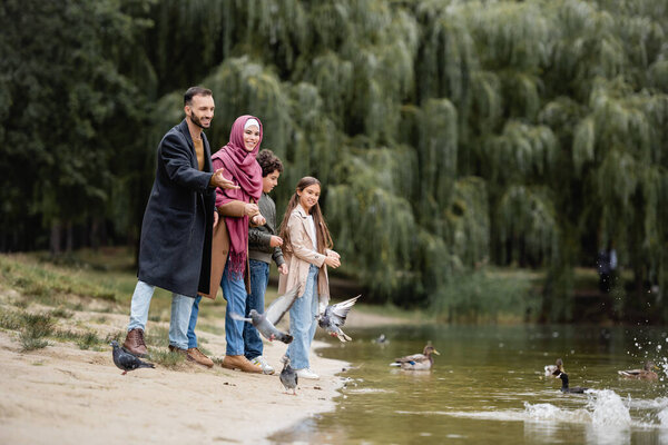 Cheerful muslim man pointing at birds on lake near family in park 