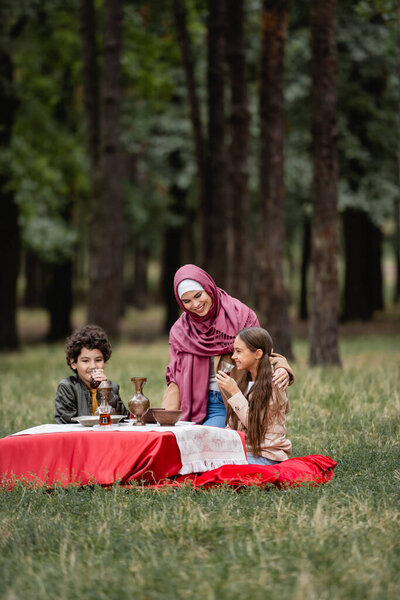 Smiling kids holing tea in glasses near muslim mother in hijab in park 