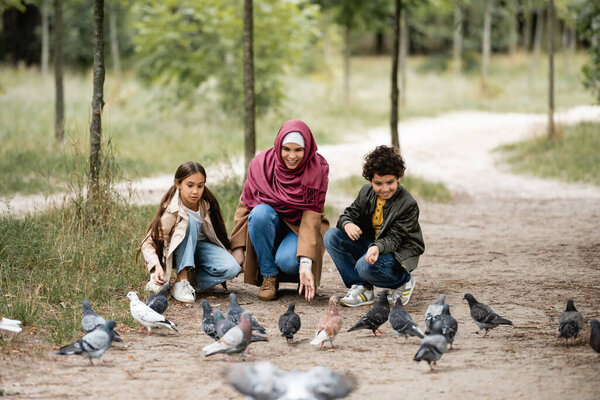 Positive arabian mother and children feeding blurred doves in park 
