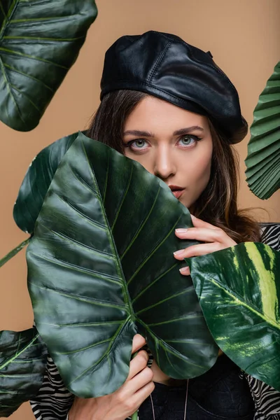 身穿黑色贝雷帽的年轻女子 穿过被米黄色隔离的绿色热带树叶看着相机 — 图库照片