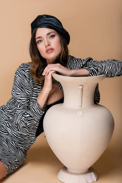 model in zebra print outfit and black beret posing near clay vase on beige