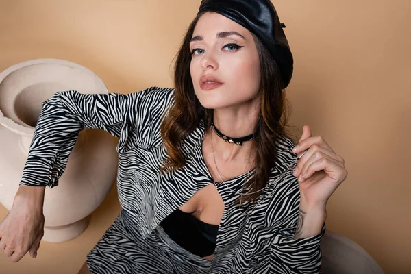 high angle view of model with tattoo in zebra print outfit and leather beret posing near clay vases on beige