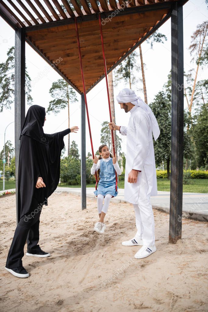 Arabian parents in traditional clothes standing near daughter on swing in park 