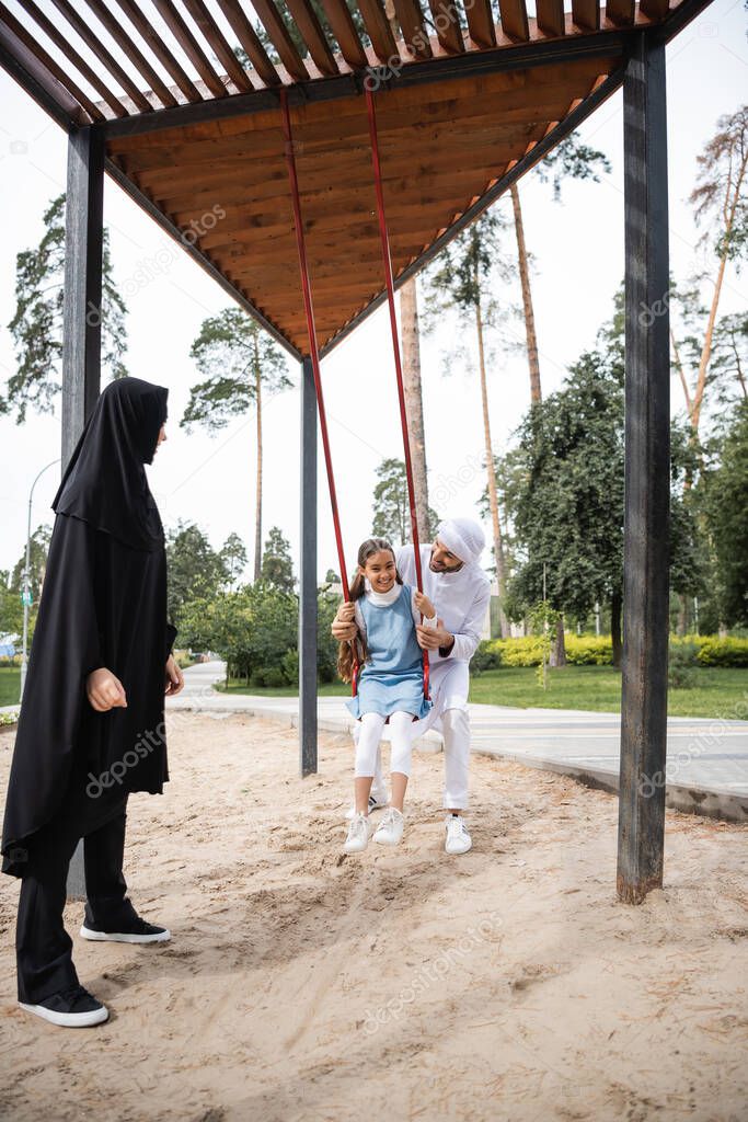 Muslim father spending time with daughter on swing and wife in park 