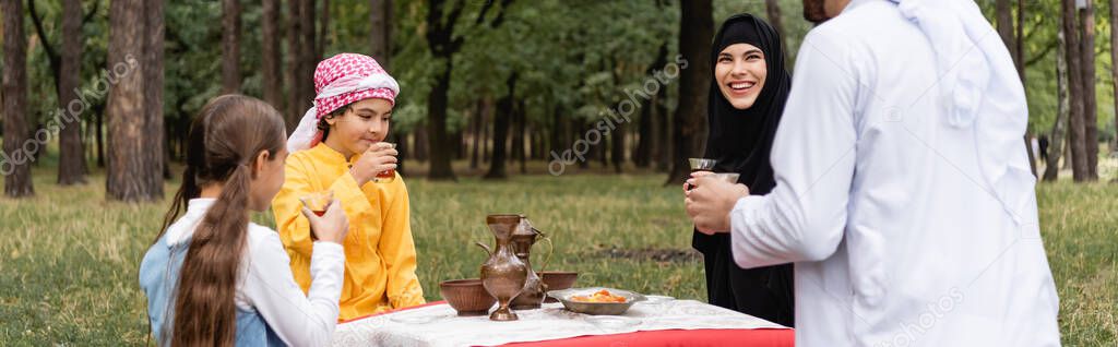 Smiling arabian woman sitting near husband, kids and tea in park, banner 