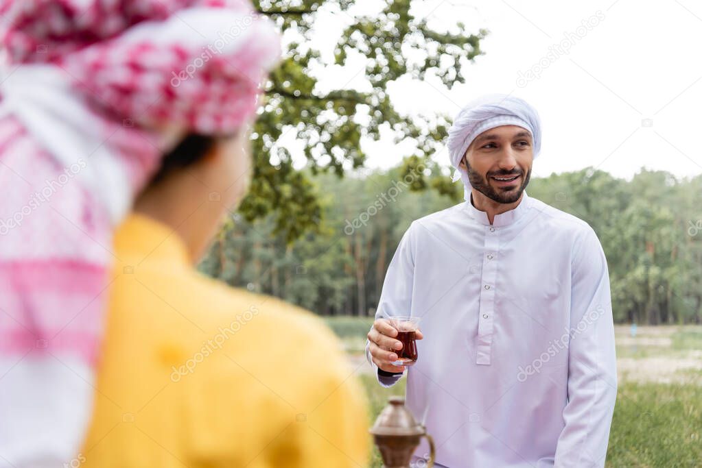 Smiling muslim man holding glass of tea near blurred son outdoors 