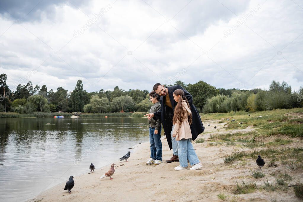 Muslim father pointing at lake near kids and birds in park 