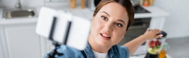 Yüksek açılı, gülümseyen, iri yarı bir kadının yanmış blender ve afiş yanında bulanık akıllı telefon kullanmasının görüntüsü. 