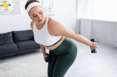 Plus size sportswoman working out with dumbbells in living room  clipart