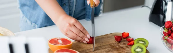 Cropped view of woman cutting fruits near blurred cellphone in kitchen, banner