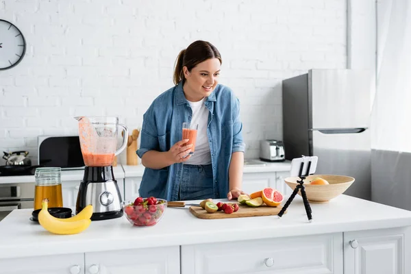 Wesoły Rozmiar Kobieta Trzyma Szklankę Smoothie Pobliżu Owoców Smartfona Kuchni — Zdjęcie stockowe