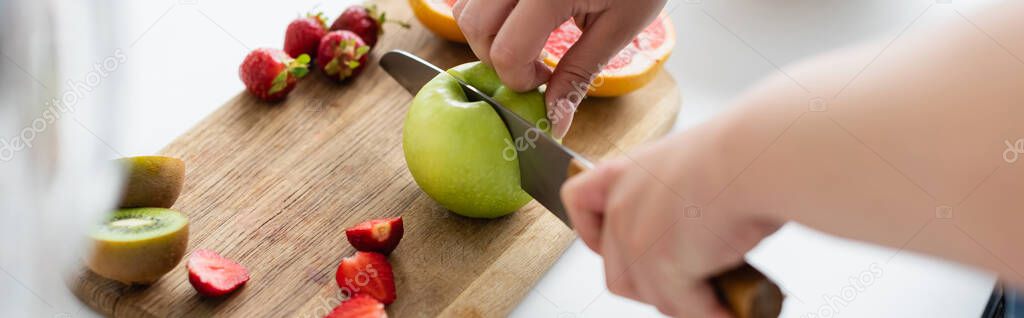 Cropped view of young woman cutting apple near fruits on cutting board in kitchen, banner 
