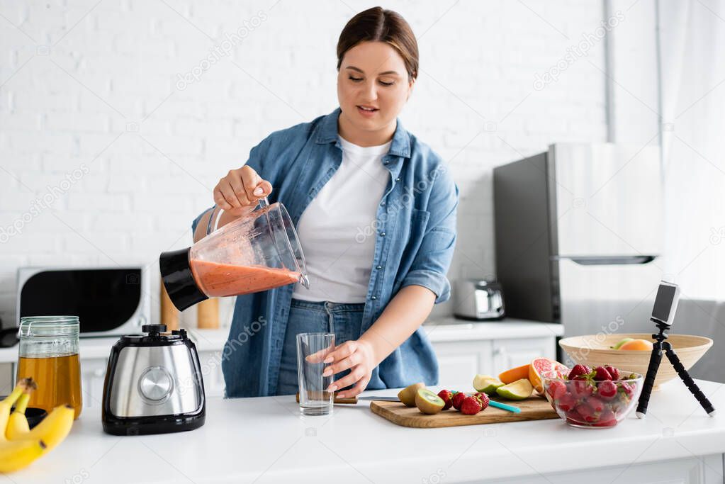 Young woman with overweight pouring fruit smoothie near smartphone in kitchen 