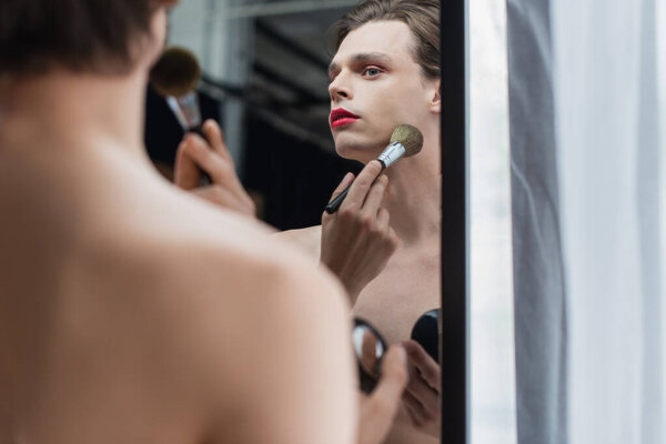 young transgender man applying face powder on neck near mirror 