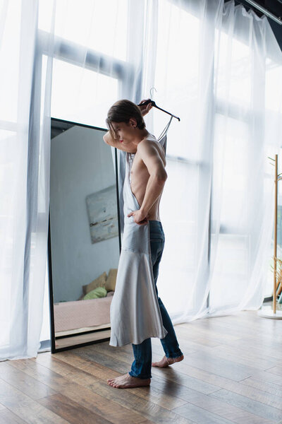 полная длина молодых трансгендерных мужчин с макияжем держа вешалку с скользящим платьем возле зеркала дома