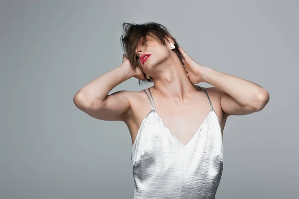 young transsexual man with makeup and earring posing in slip dress isolated on gray