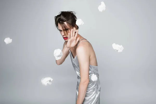 scared transgender man in slip dress protecting from crumpled paper attack on gray