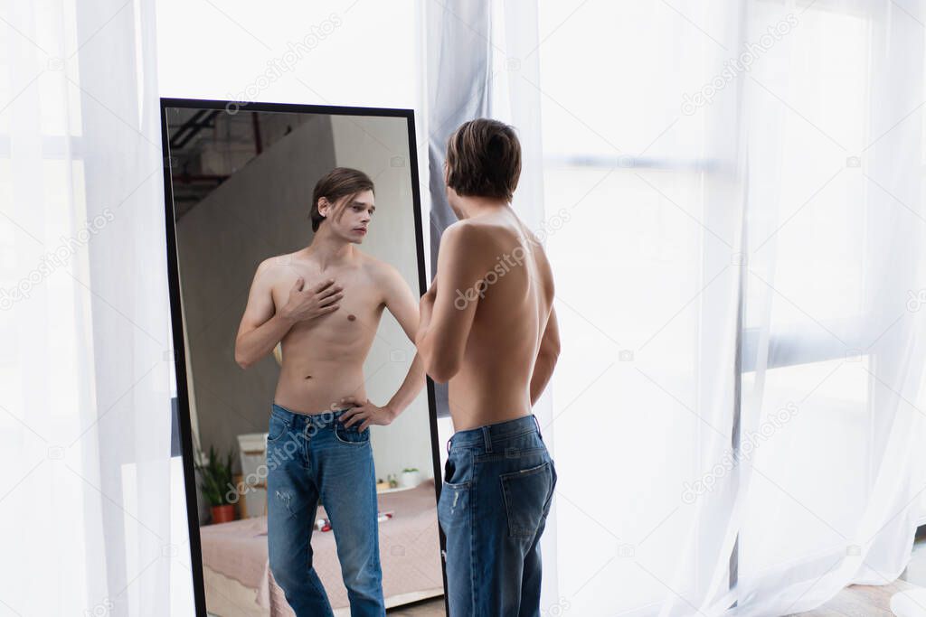 shirtless transgender young man in jeans standing with hand on hip while looking at mirror 